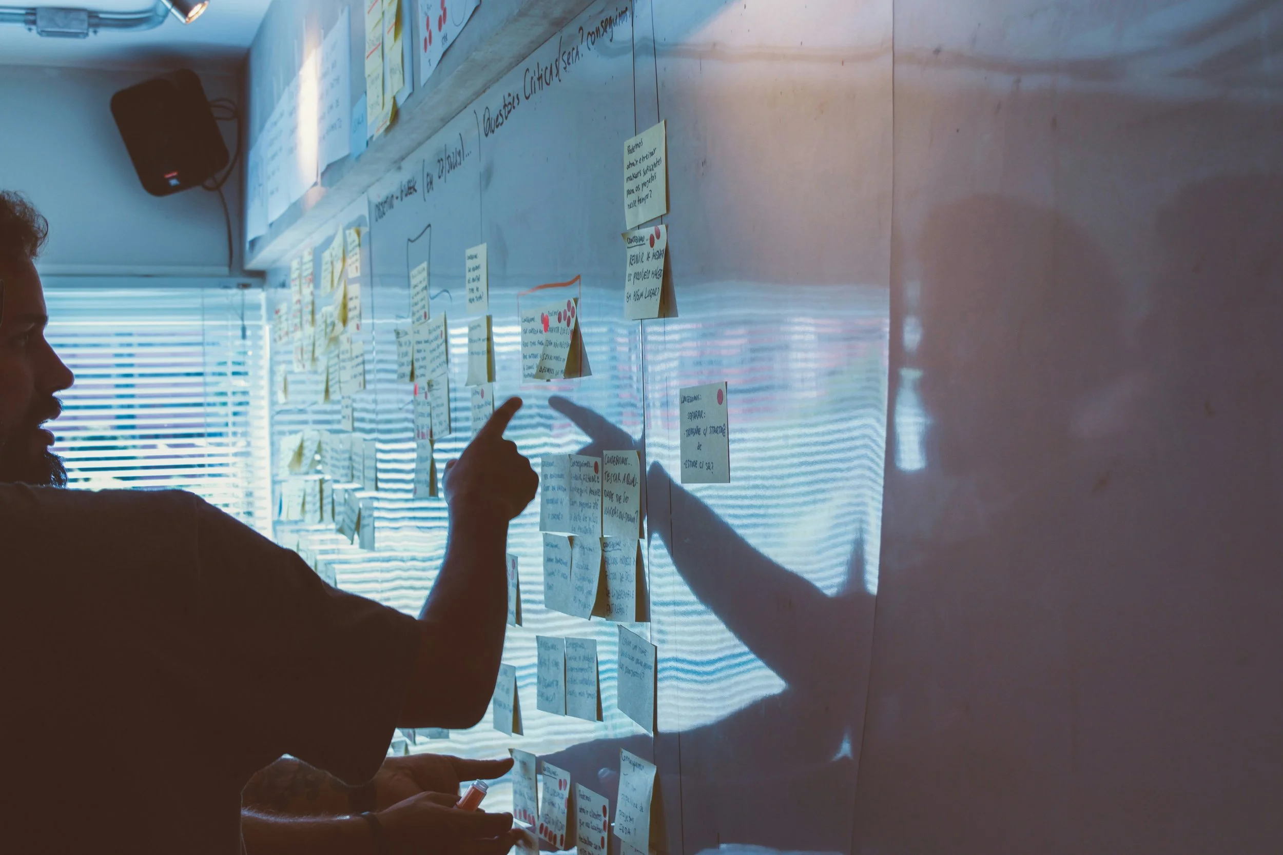 man in a dim room pointing at whiteboard covered in stickynotes